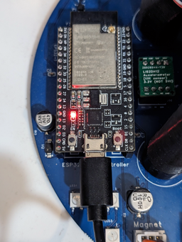 An image of the ESP32 module mounted in the 2023 circuit board with a black USB cable plugged into the bottom of the ESP32 and the module's power LED shining red